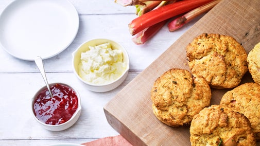 Scones on a wooden serving plate next to jam, cream and rhubarb